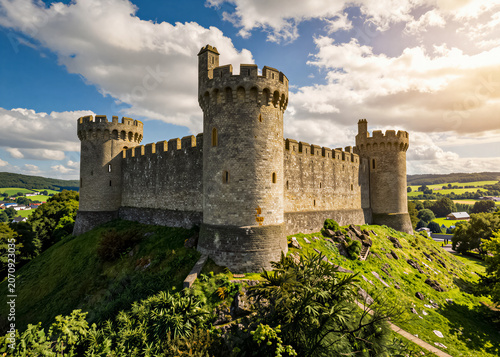 An ancient stone castle with high towers and walls, perched atop a lush green hill. The sun breaks through picturesque clouds, illuminating the historic fortress overlooking the lush valley
