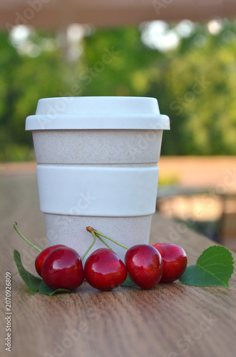 Closeup white -beije paper coffee cup with lid and fresh red cherries with green leaves arranged   on wooden table background. Morning coffee to go and cherry fruit , summer dessert  concept 