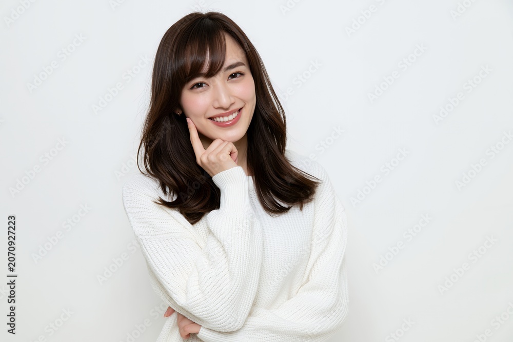 Fototapeta premium 白いセーターを着た笑顔の若い日本人女性が頬に指を当てる