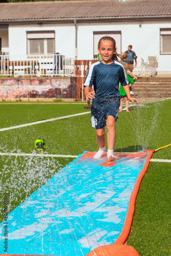 Fototapeta premium Child running on a colorful water slide with splashes of water in a sunny outdoor setting, surrounded by green grass and a residential background
