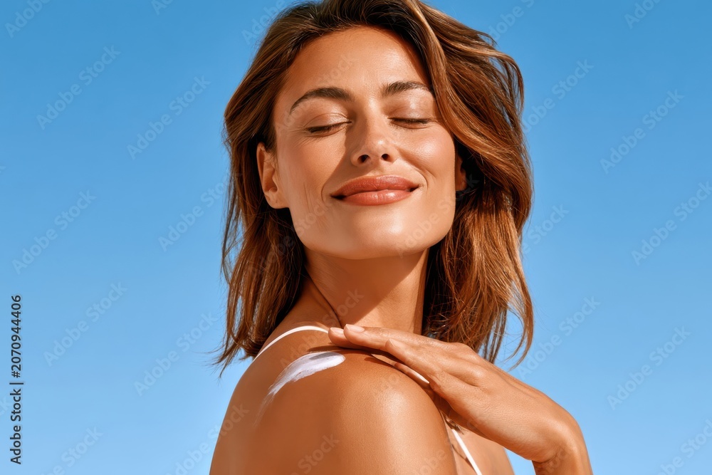 Fototapeta premium Young woman applying sunscreen on shoulder, smiling with eyes closed under bright summer sun