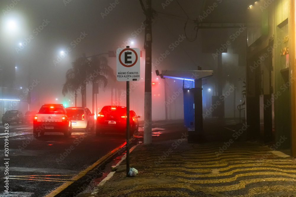 Naklejka premium A car drives through thick fog on an urban street under traffic lights at night in downtown Marilia, Sao Paulo state,