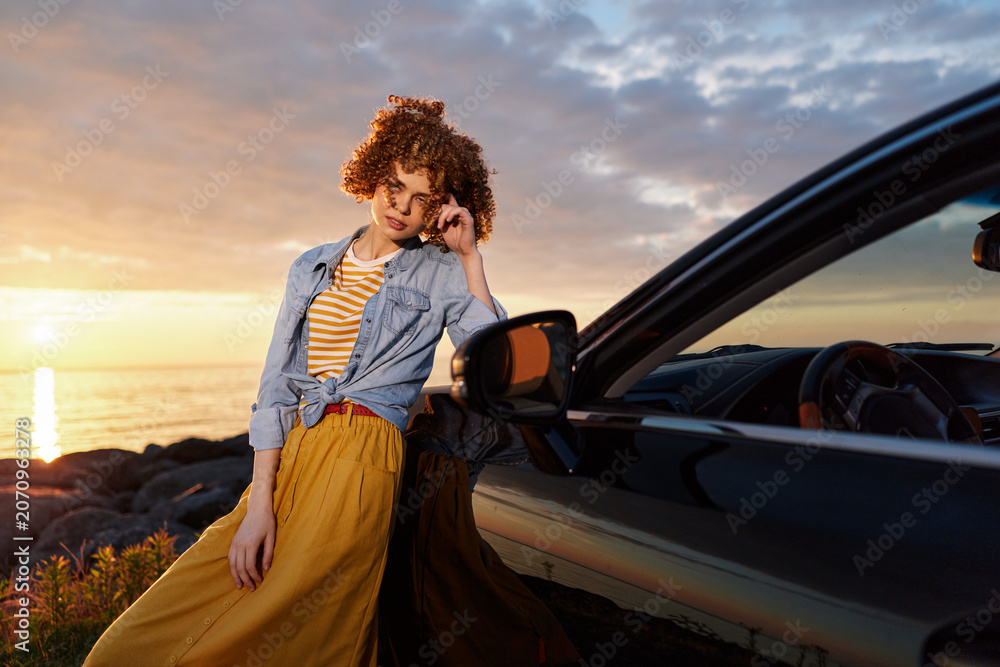 Fototapeta premium A woman with curly hair rests against a sleek car at sunset, wearing a striped tee and denim jacket, showcasing a casual, carefree lifestyle and travel vibe.