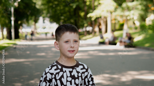 Portrait of boy standing on tree-lined park pathway in daytime, wearing patterned black and white shirt, with neutral expression and soft dappled light