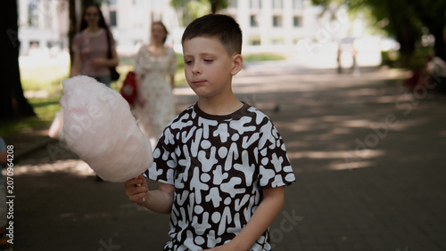 Young boy carries fluffy pink cotton candy on stick while walking through leafy urban park path with dappled sunlight and soft blurred background