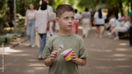 Boy in green shirt holding colorful ice cream cup and spoon while standing on shaded park walkway; soft daylight filtering through trees creates dappled