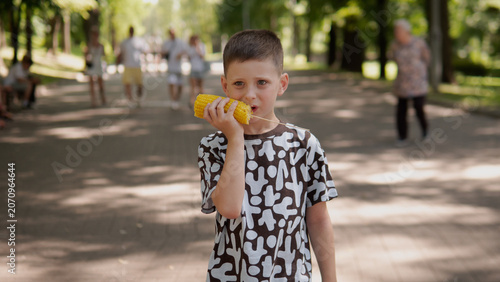 Young boy holding corn on cob to mouth while standing on paved park walkway surrounded by tall trees and dappled sunlight, casual patterned t-shirt, relaxed