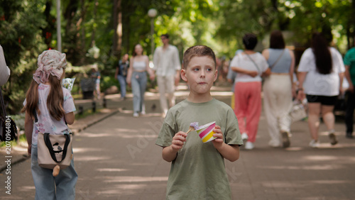 Boy in olive green t-shirt holds colorful paper airplane while standing on shaded pedestrian path in leafy urban park; blurred walkers and couples move behind