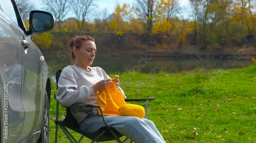 Woman knitting outdoors during an autumn road trip. Relaxed woman sitting on a camping chair next to her car, enjoying a peaceful afternoon knitting with yellow yarn in a autumn park by the river