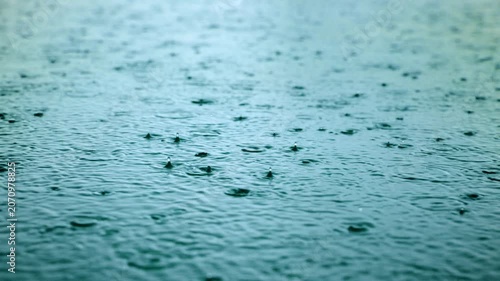 Rain falling on water creates mesmerizing ripples and splashes in slow motion. Teal-toned water surface captures raindrops bouncing and forming concentric circles in a calming natural pattern.