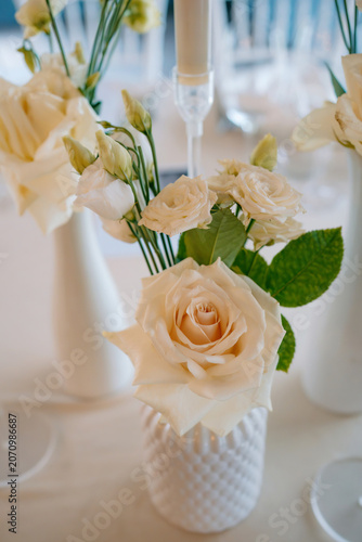 Cute wedding table centerpiece featuring ivory roses, delicate flower buds and green foliage arranged in a decorative white vase, sophisticated floral display for a wedding reception or special event