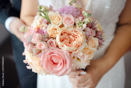 Young bride wearing a beautiful tulle white wedding gown while holding a lush bouquet of pink and cream roses, peonies and delicate seasonal flowers in a romantic pose holding arms with her groom