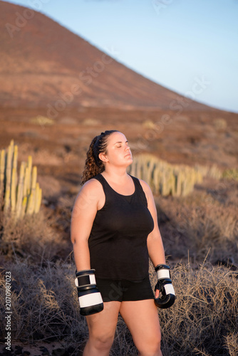 Powerful young woman wearing a black athletic outfit and boxing gloves while standing with her eyes closed surrounded by arid vegetation during golden hour, enjoying a break in her fitness routine