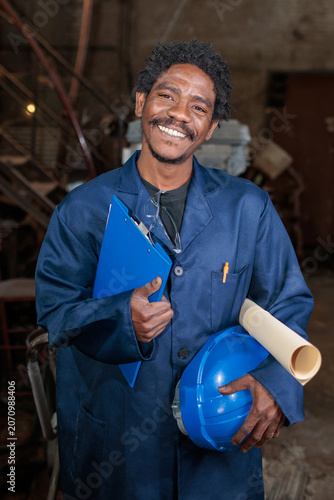African worker in a factory holds a clipboard and a helmet while smiling at the camera