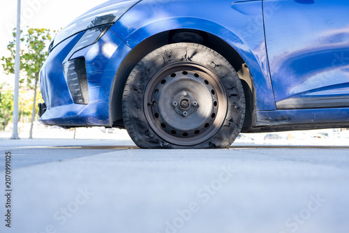 Flat Tire on Blue Car Parked on Street. A close up view of a deflated vehicle tire on a sunny day depicting an automotive emergency or a mechanical failure requiring repair and roadside assistance for