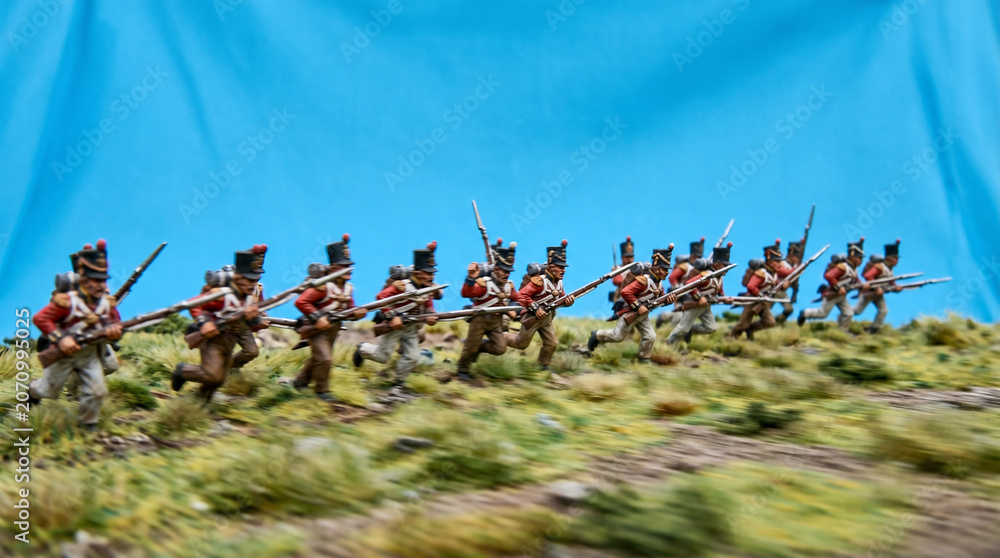 Fototapeta premium March of Time: A regiment of 19th-century soldiers in red uniforms advance across a grassy incline, muskets at the ready, evoking a sense of historical military action and discipline.