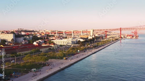 Aerial drone shot of 25 de Abril suspension bridge above Tagus River in Lisbon Portugal. Scenic waterfront cityscape with riverside promenade and urban skyline at sunset