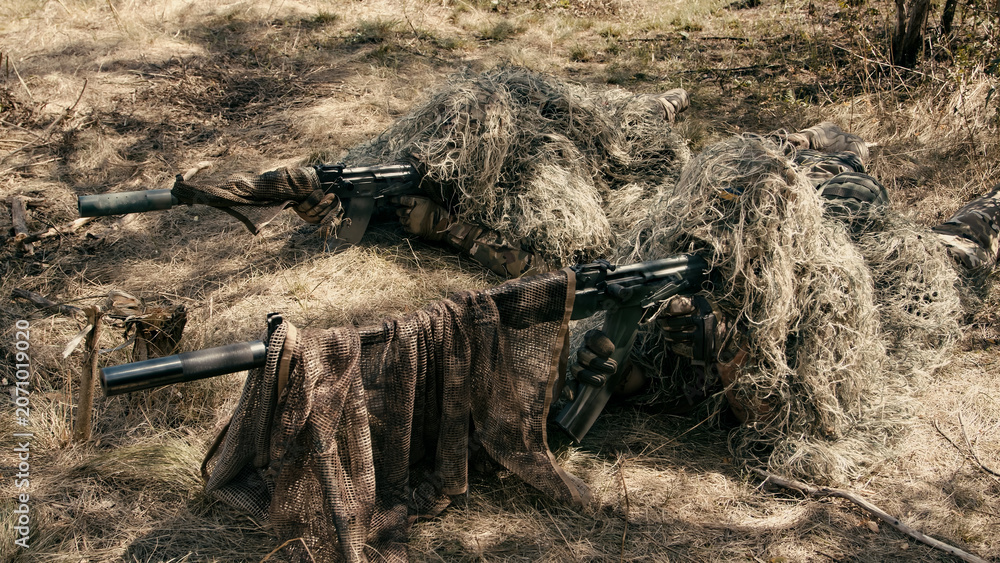 Naklejka premium Sniper team camouflaged with ghillie suits lying on the ground in a forested area, rifles positioned for a tactical mission in a natural environment