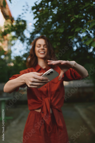 Bild auf Leinwand Smiling woman in a red dress enjoying a casual outdoor moment, taking a selfie in a garden setting