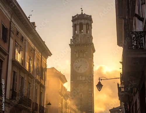 A tall, stone clock tower rises amid buildings. Sunlight streams, creating a warm, hazy atmosphere. Street lamps hang
