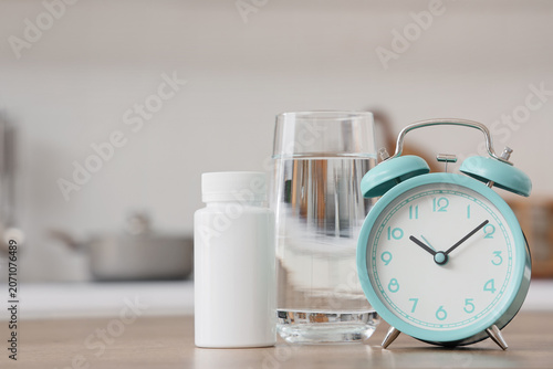 Fotografie Alarm clock with pill bottle and glass of water on table in kitchen, closeup