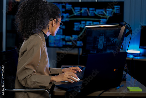 Black female developer uses a tablet to annotate a data visualization on a large monitor in a dark server operations center.