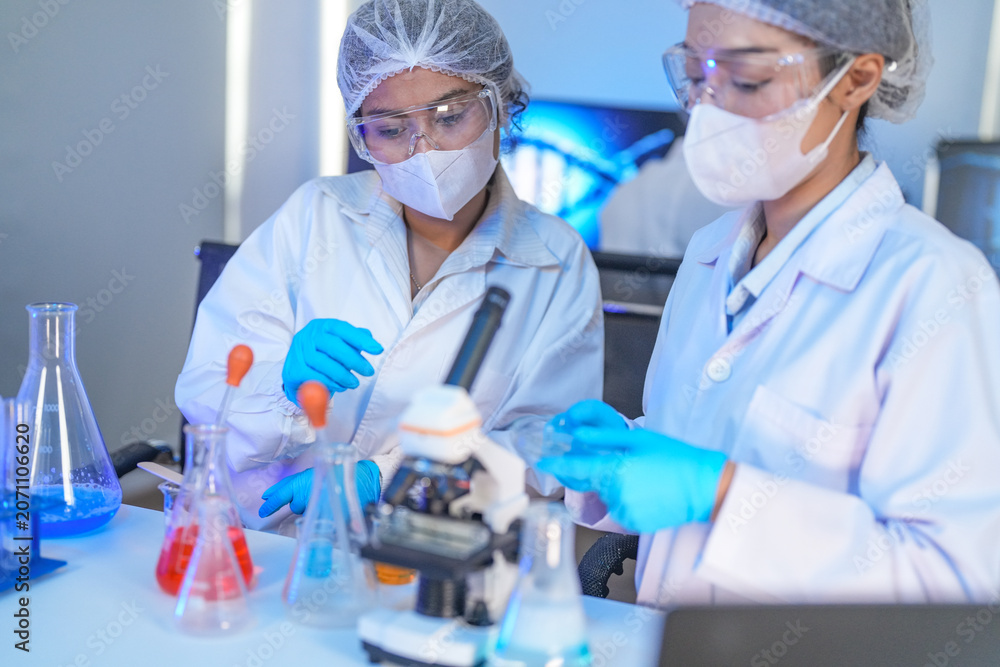 Fototapeta premium Scientists in lab coats and masks examine samples with a microscope and test tubes in a modern research laboratory.