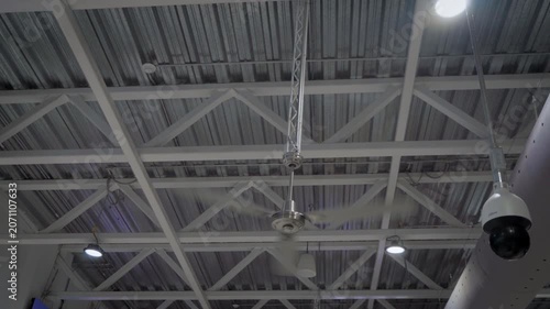 Handheld shot captures a large industrial fan in motion inside a warehouse, its spinning blades filling the frame against a gray ceiling structure background in a clean neutral industrial setting