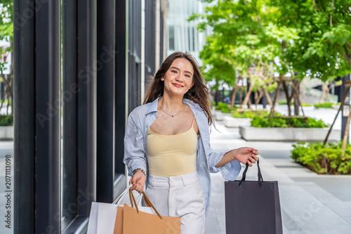 Happy young Asian woman walking on a modern city street holding shopping bags. She smiles and looks at store windows enjoying a weekend shopping trip. Fashionable lifestyle and retail therapy.