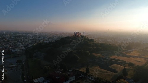Aerial drone moves cinematically close to the Church of Our Lady of Remedies atop the Cholula pyramid at dawn, with the town and Puebla city spreading across the background
