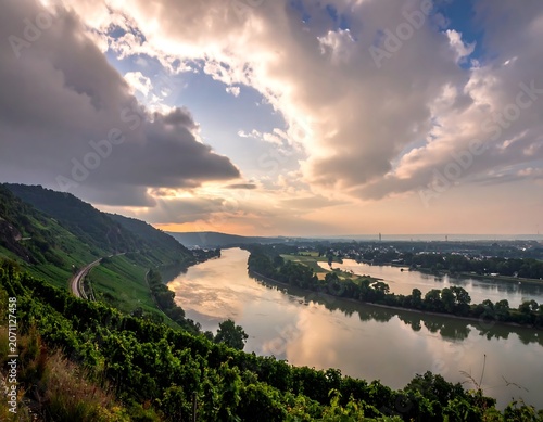 A sweeping vista showcases a meandering river, shadowed by lush greenery. The sky, a tapestry of clouds, filters sunlight over the landscape