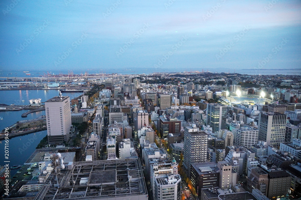Obraz premium 夜明け前の都市風景。海岸線に広がる港湾と高層ビル群、照明が輝くスタジアムが夜空に映る。