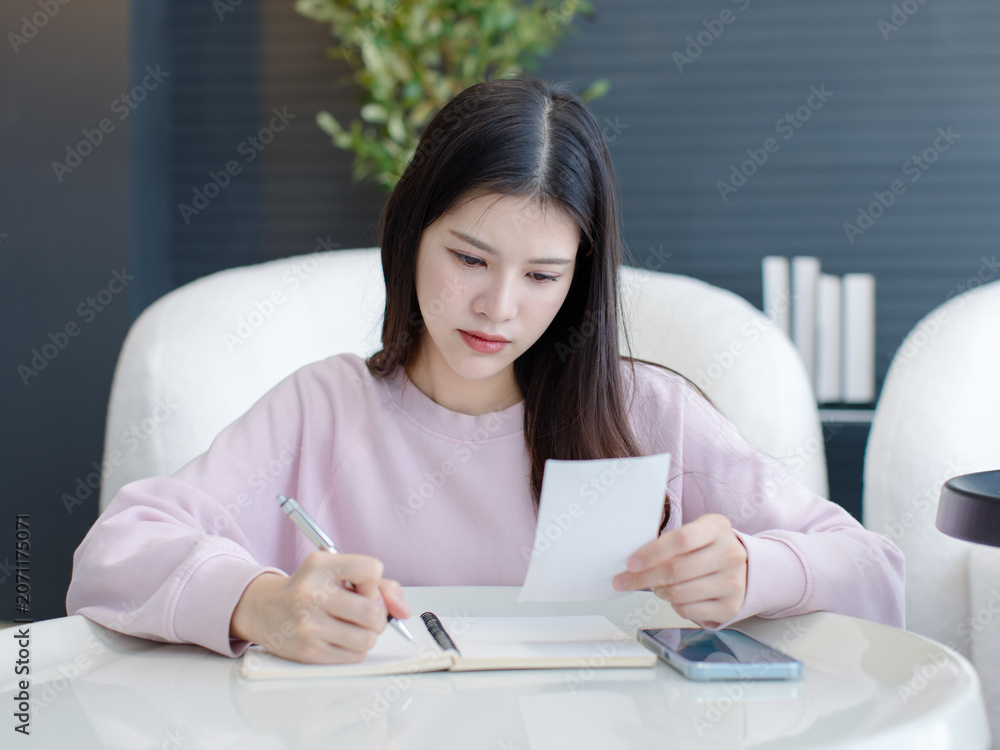 Naklejka premium Asian happy beautiful woman working writing record information on paper sitting on modern living room ,lifestyle enjoy wellbeing healthy young person relaxed mood peaceful security wellness at home.