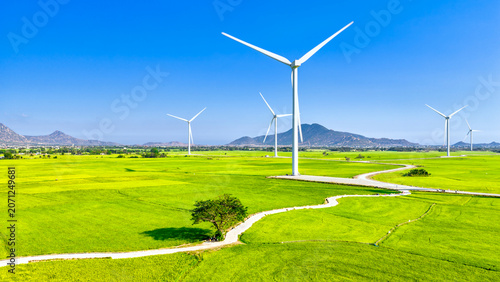 Bright sunny day at the Ninh Thuan wind farm, Vietnam, featuring modern wind turbines rising above vast green agricultural fields. The clear blue sky, winding rural roads