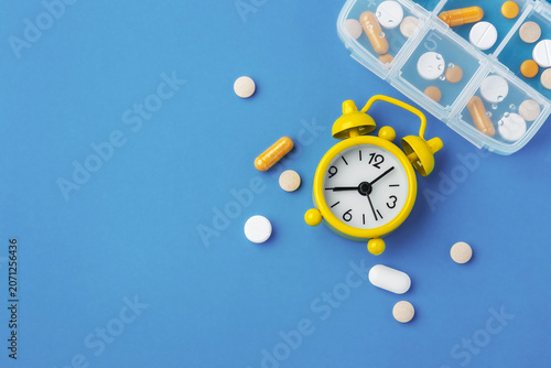 Medicine pills, tablets and capsules with yellow alarm clock and pill box on blue background, top view