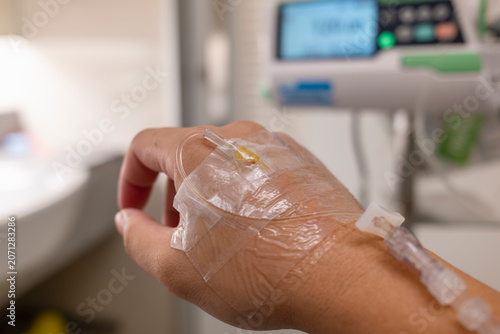 Patient hand with IV drip fluid line close up in hospital room with blurred digital infusion pump monitor background. Healthcare medical insurance policy, illness treatment, and care concept.