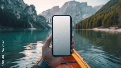 A person holds a smartphone with a blank screen while kayaking on a serene mountain lake surrounded by forested peaks