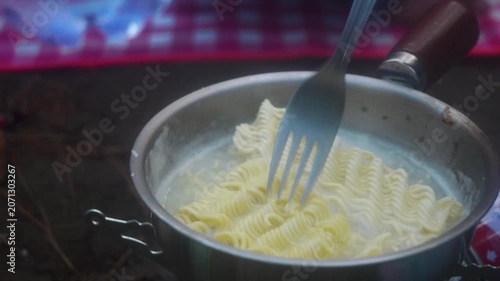 Close up of noodles being stirred in a small pot with a fork during outdoor cooking, preparing a quick meal while camping in nature and enjoying a simple culinary experience