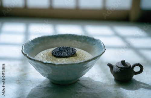 A shallow handmade celadon ceramic bowl of smooth rice pudding dusted with fine matcha powder a single black sesame tuile resting on top placed on a weathered washi paper surface beside a tiny clay 