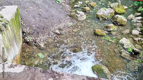 Clear water flows through a shallow rocky stream beside a weathered concrete structure. Moss covered stones, gentle currents, and natural textures create a peaceful countryside scene