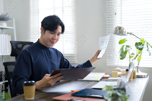 Canvas-taulu Young Asian professional reviewing business documents at bright home office desk