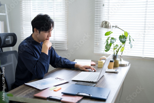 Obraz na płótnie Young Asian businessman deep in thought working on laptop with financial charts