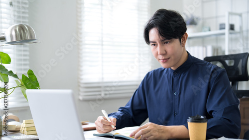Bilde på lerret Young Asian professional writing notes in office while working on laptop during
