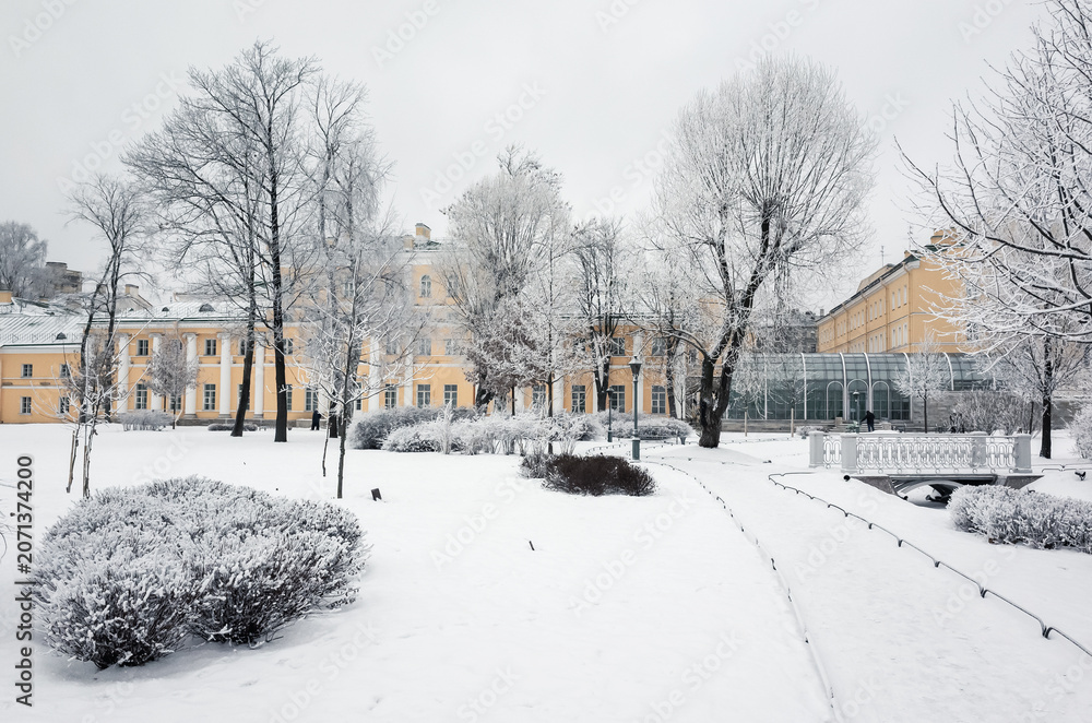 Fototapeta premium A scenic snow-covered park featuring frosted trees and a historic yellow buildings, evoking a serene and magical winter atmosphere in an urban setting of The Polish Garden, Saint-Petersburg, Russia