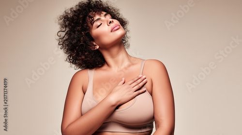 Curly-haired woman gently touching her chest, embodying self-love, body awareness and wellness against beige background. Perfect for healthcare, self-care and women's wellbeing campaigns.