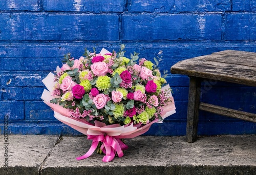 Vibrant flower bouquet with pink carnations and roses in bright wrapping paper next to a wooden bench against a blue brick wall.