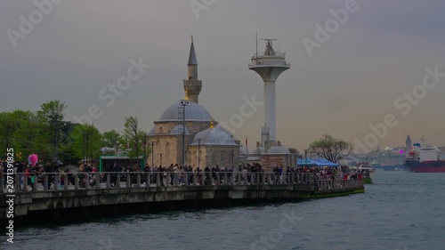 People walking along the Uskudar promenade in Istanbul featuring a beautiful mosque and white lighthouse by the Bosphorus sea during a calm evening sunset