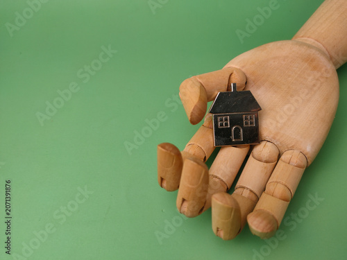 A silver house model rests on the open palm of an articulated wooden mannequin hand against a green background