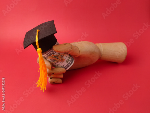A wooden mannequin hand holds a roll of dollar bills wearing a miniature graduation cap with a yellow tassel