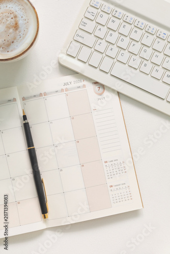 Top view of a clean workspace with monthly planner, laptop keyboard, coffee cup and pen on white desk. Productivity, scheduling, remote work and business organization concept with copy space.